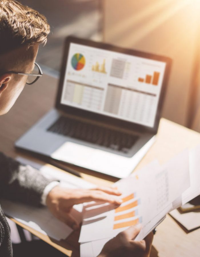 Young finance market analyst in eyeglasses working at sunny office on laptop while sitting at wooden table.Businessman analyze document in his hands.Graphs and diagramm on notebook screen.Blurred